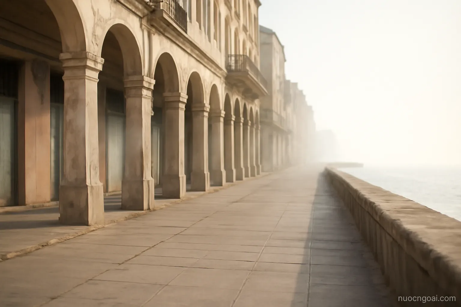 Low colonnaded streets of weathered pastel buildings meeting a long sea wall under bright haze
