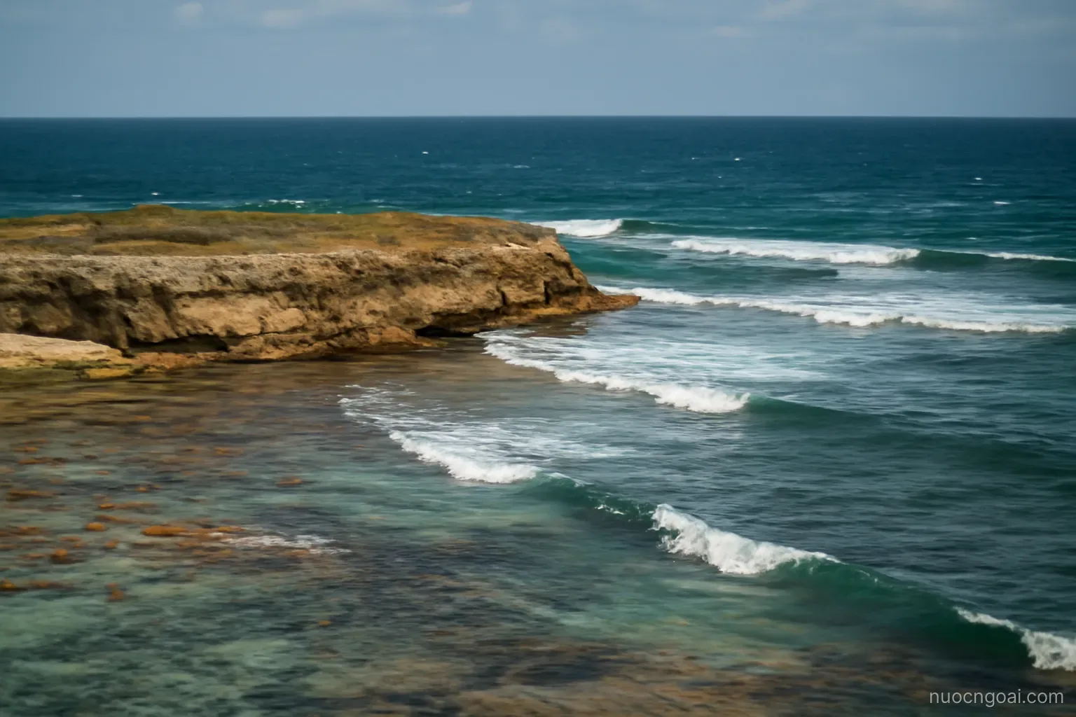 A flat coral island edge of low pale cliffs and reef flats with open Atlantic swell arriving from the east