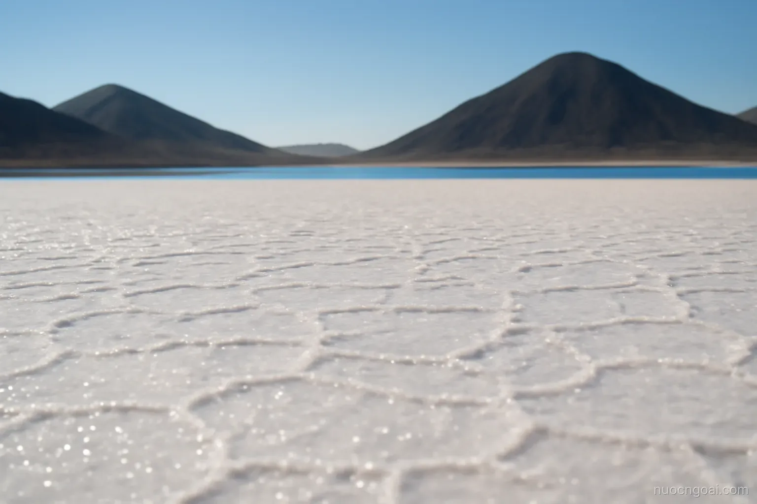 A salt flat shimmering under midday heat with dark volcanic hills rising behind a narrow strait