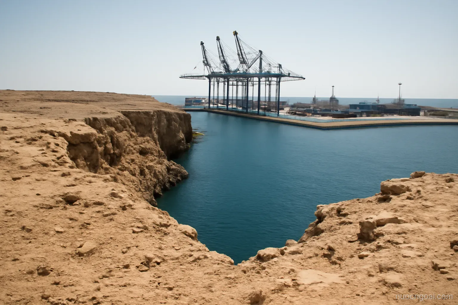 A dry rocky plateau dropping to a narrow gulf, container cranes on a single quay in hard midday light