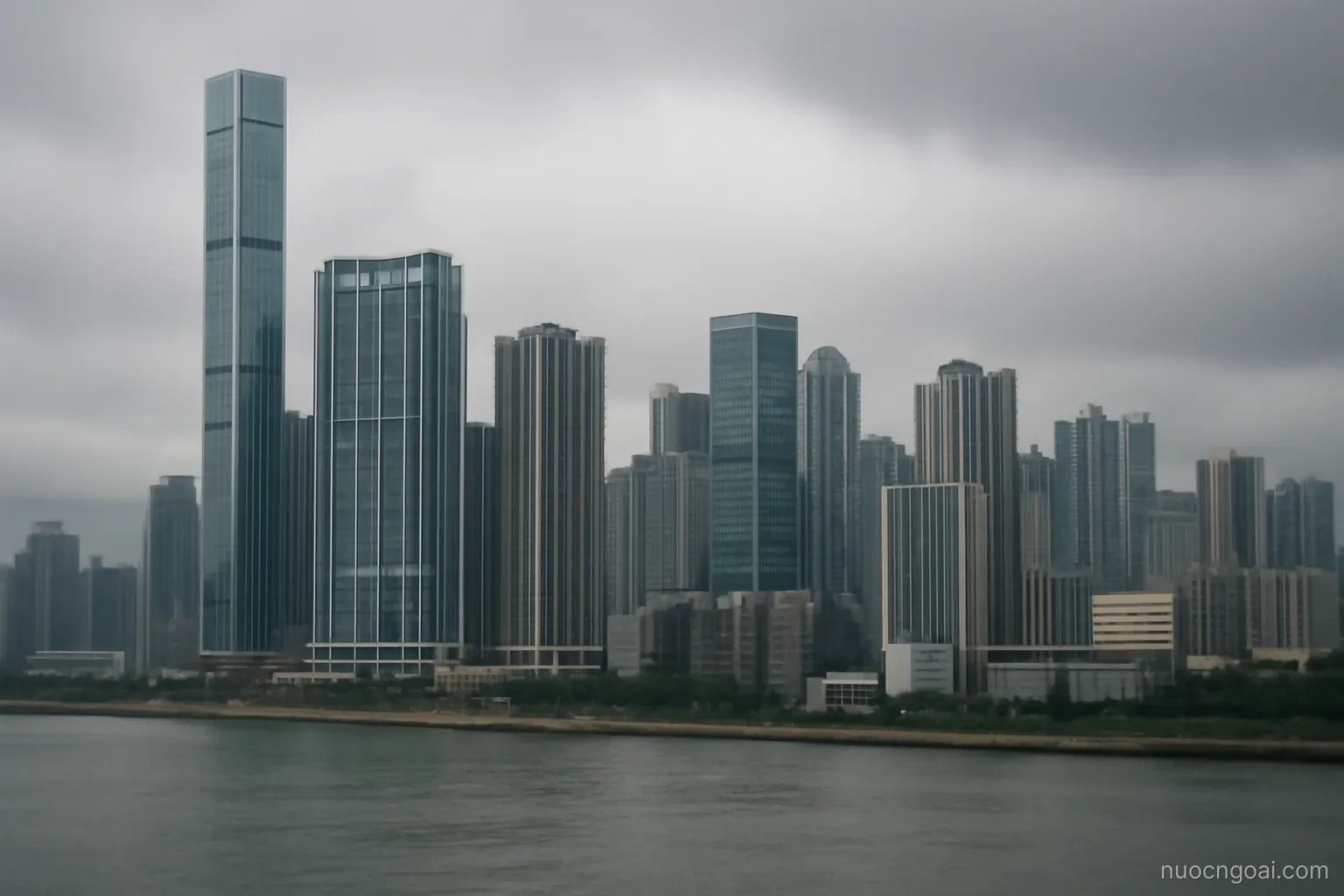 A dense waterfront of tall towers on reclaimed land beside a grey estuary under low cloud
