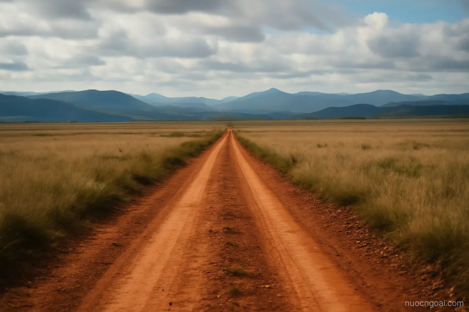A red-earth road running for miles across open highland grassland towards distant blue ridges under high cloud