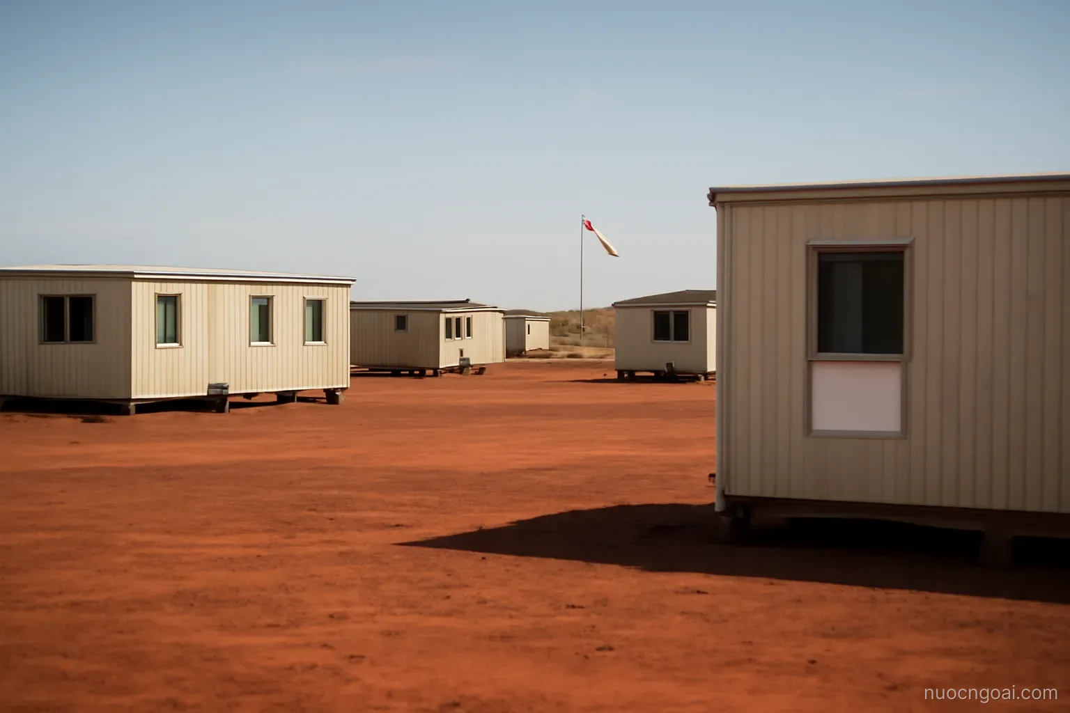 A remote mining camp of low prefabricated blocks on flat red laterite ground with an airstrip windsock in the distance