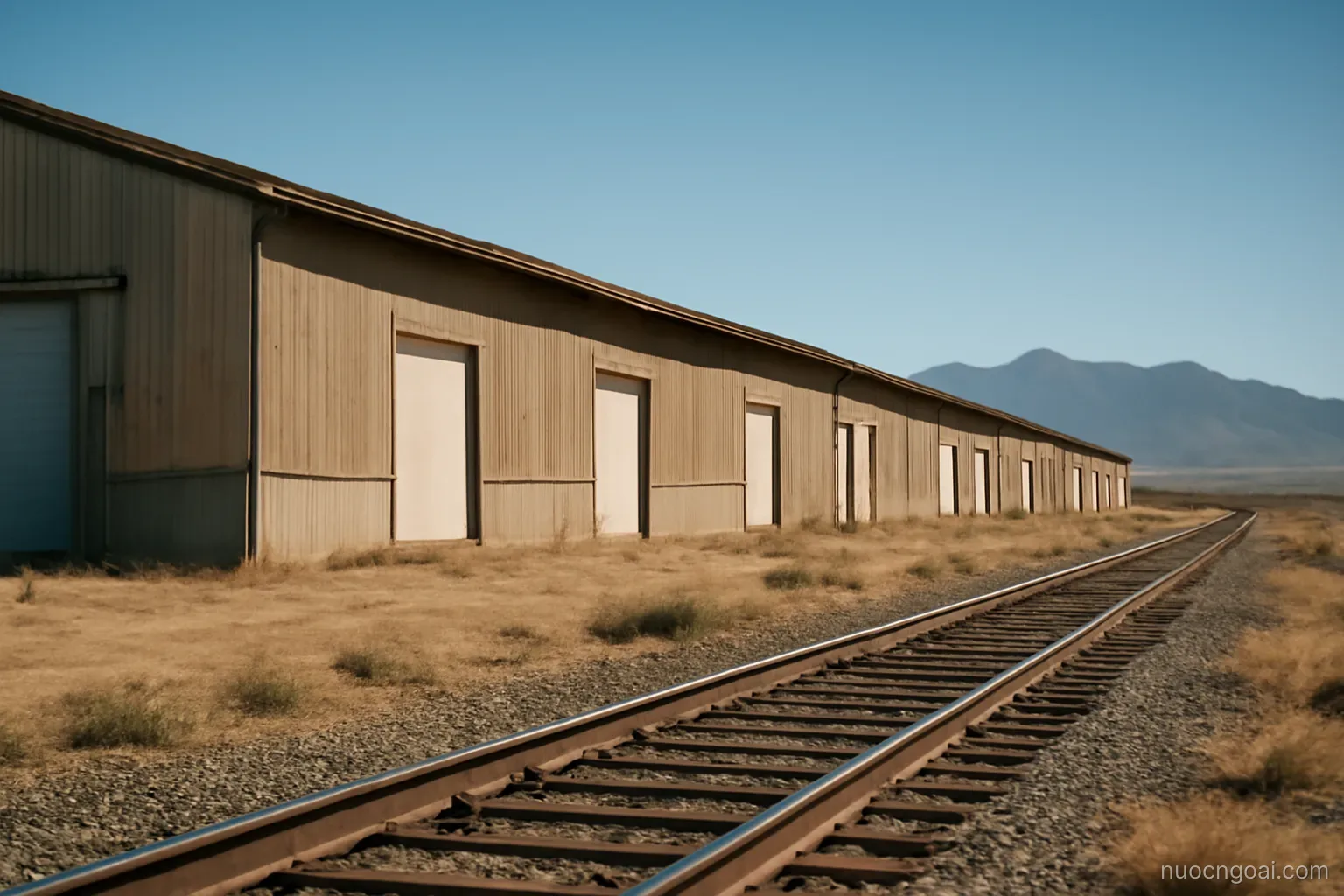 Long low factory sheds beside a rail line on dry high plateau, distant blue sierra under clear sky