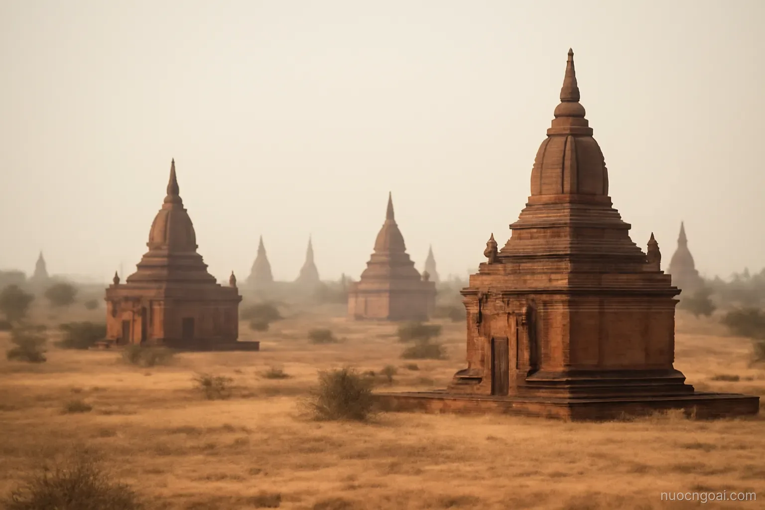 A broad dry plain scattered with brick stupas fading into dust haze under a hot white sky