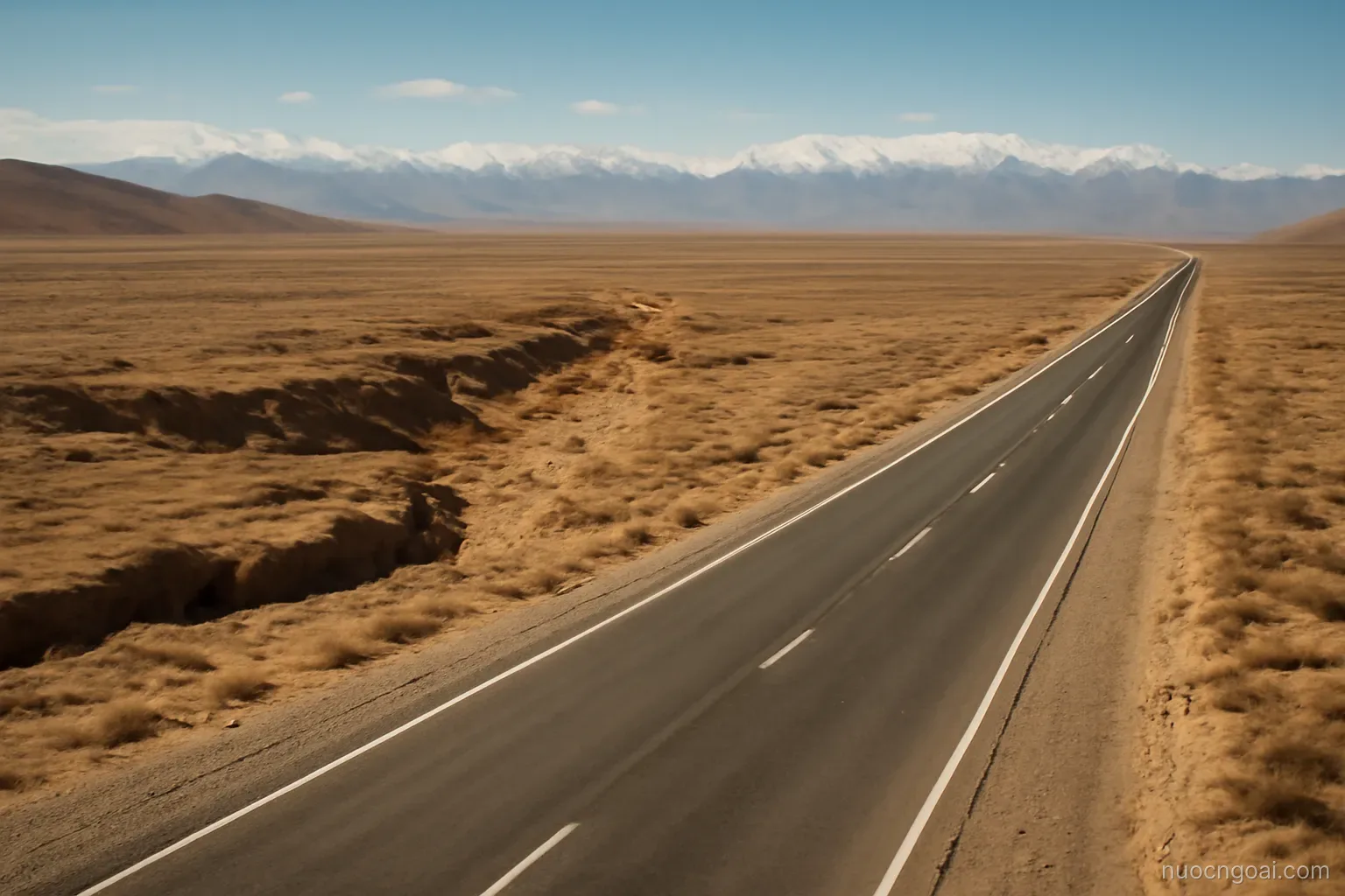 Arid plateau cut by a dry riverbed, distant snow ridges and a single paved road under hard light