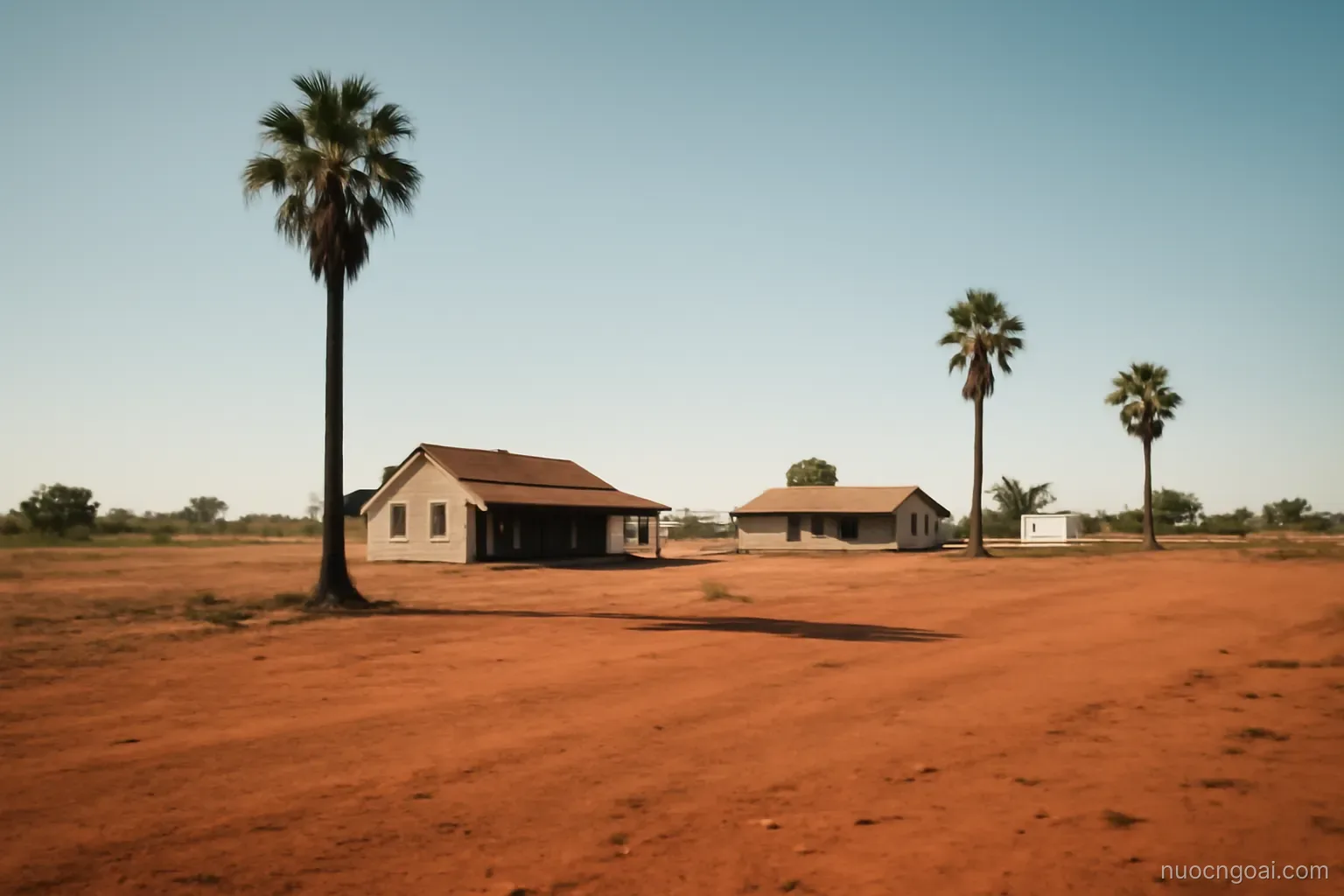 Flat red-earth countryside with scattered palms and low ranch buildings under a hot bright sky