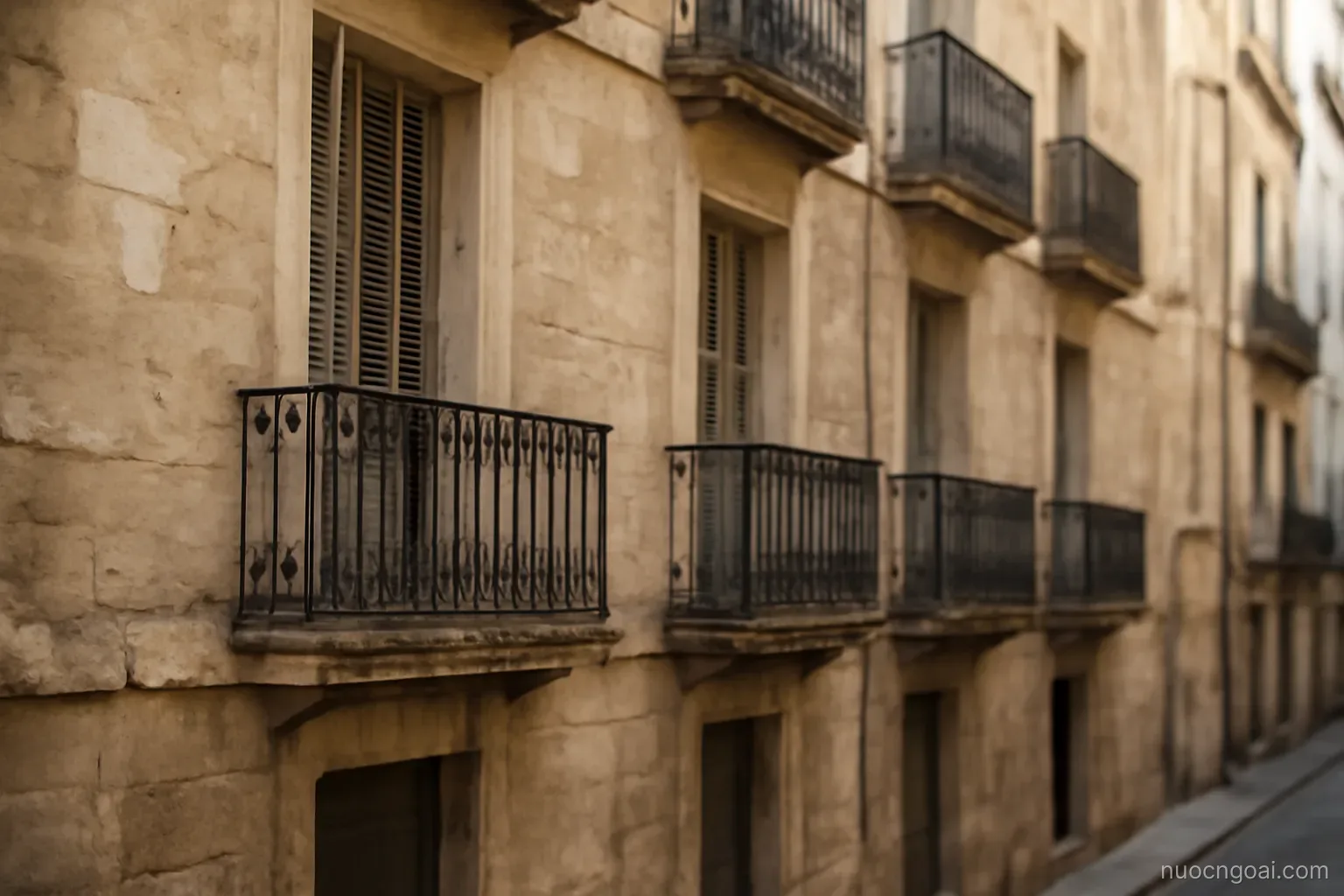 A stone apartment façade with wrought-iron balconies above a narrow street, shutters half closed at noon