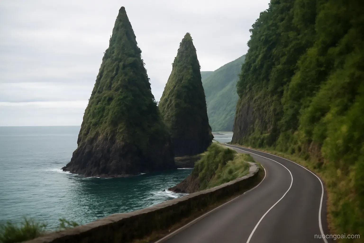 Two steep forested volcanic spires rising straight out of the sea beside a narrow winding coastal road