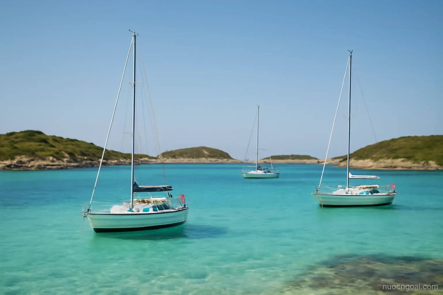 Sailing yachts anchored in a shallow turquoise bay between several low scrub-covered islets under clear sky
