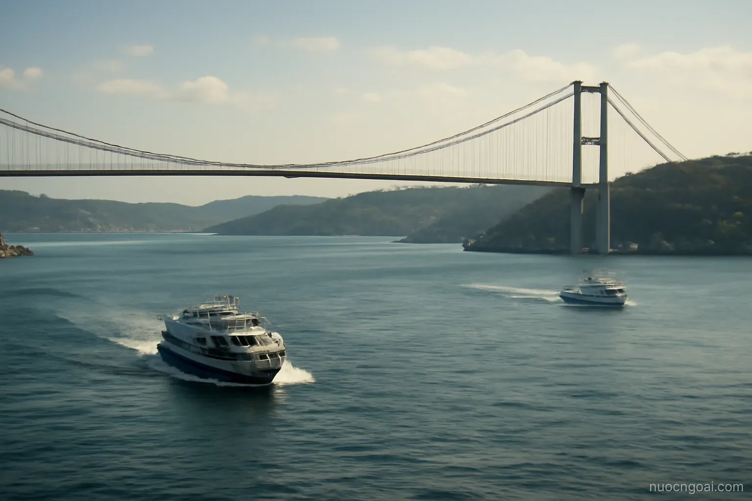 A strait crossed by a long suspension bridge, ferries cutting white wakes between two hilly shores