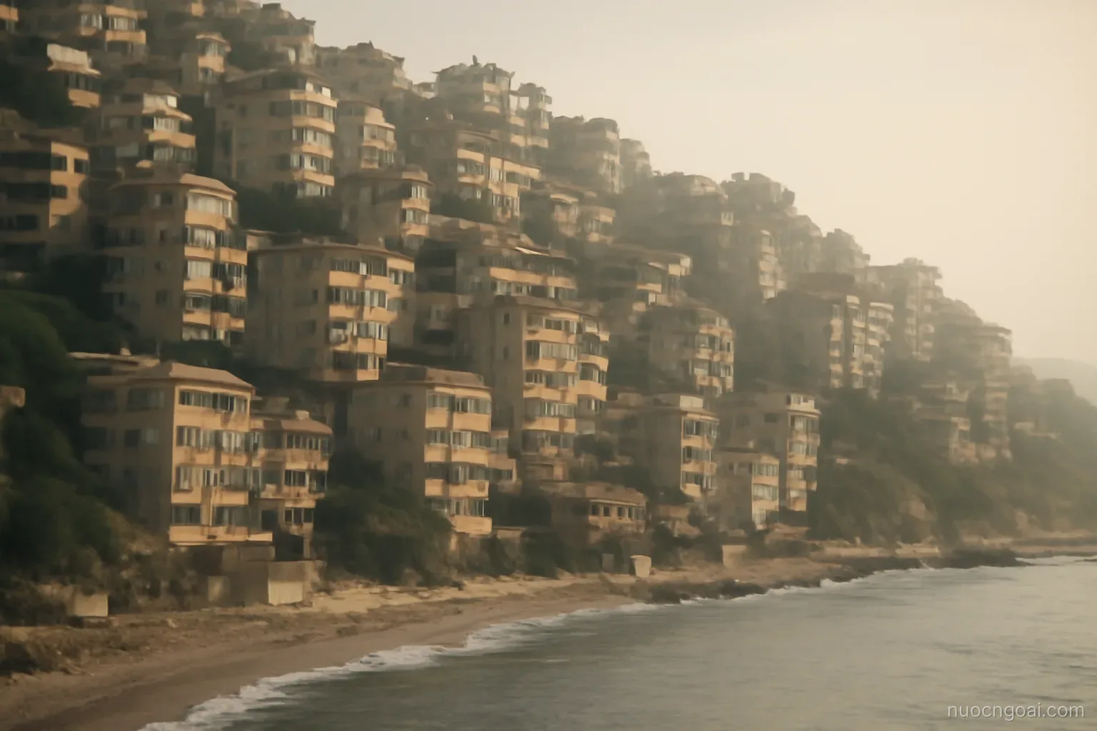Steep coastal hills of stacked apartment blocks above a narrow shoreline under hazy afternoon light