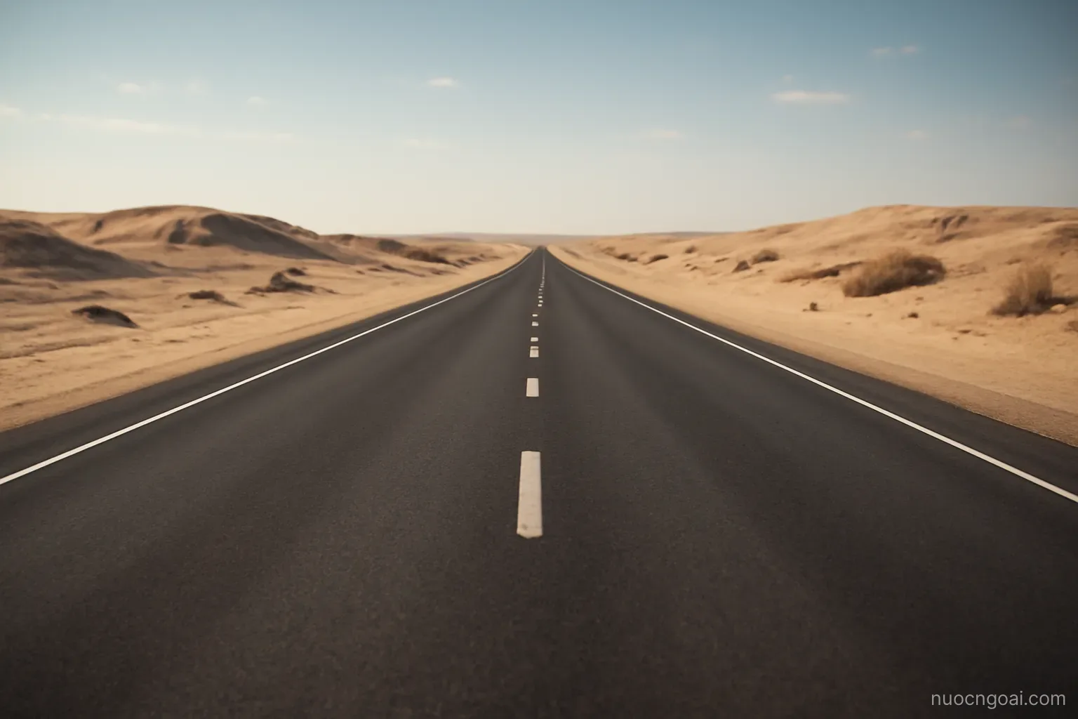 An empty four-lane desert highway running straight to the horizon between low sand ridges